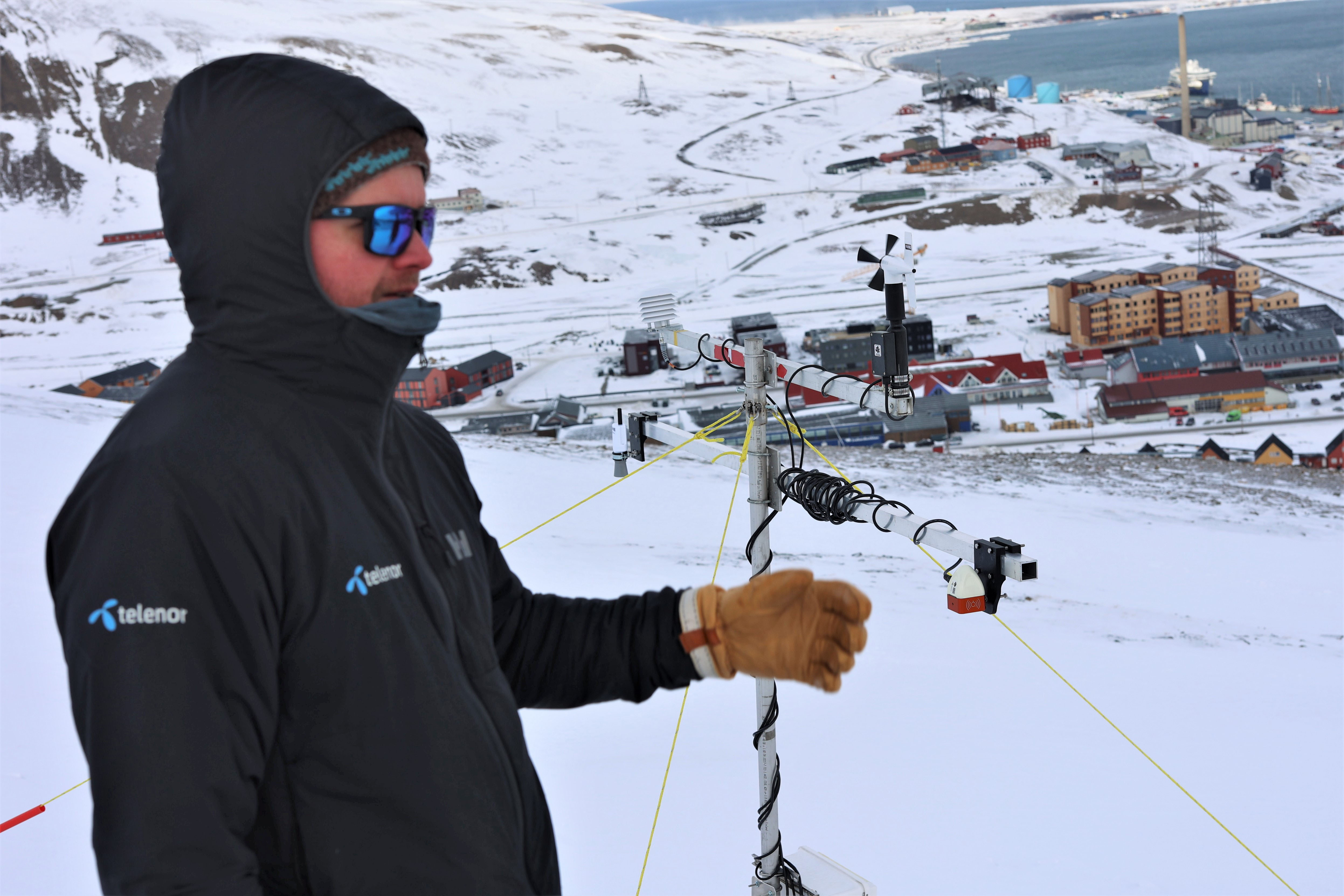 A snow sensor set up on a hillside. A man visible in the foreground and Longyearbyen in the background.